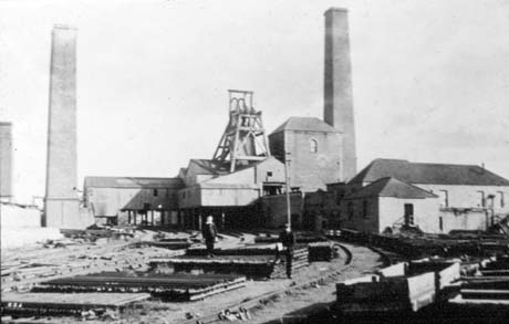 Photograph showing three chimneys, winding gear, winding house, and other buildings of a colliery; in the foreground are coal waggons on the right and piles of wood or metal in between the line on which the waggons are and another railway line; the colliery has been identified as the colliery at Station Town; a boy is standing on the piles of wood