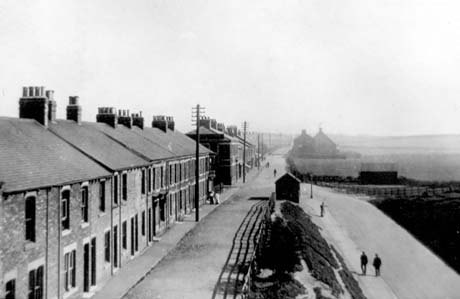 Photograph, taken from above, showing a line of terraced houses running along the left of the picture into the distance; a road is running parallel with the terraces; on the right of the photograph is a fence and, behind it, a slope leading down to a road running back towards the camera; in the distance, on the right, open fields can be seen indistinctly; the photograph has been identified as Station Town