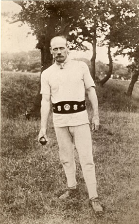 Photograph of a man dressed in a short-sleeved shirt and cloth sport trousers with a trophy belt and carrying a shiny ball in his right hand; he is standing in front of grass and trees; he has been identified as Tom Nicholson, Champion Pot-Share Bowler, Thornley