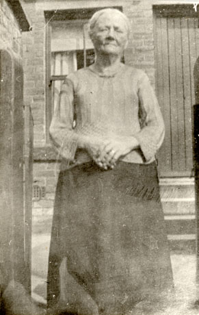 Photograph of an elderly woman standing in the gateway of a house with a window and doorway of the house behind her; she is wearing a long dark skirt and a lighter coloured jumper; she has been identified as Granny Garthwaite, Thornley