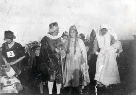 Photograph of, on the left of the picture, a woman with the number two on her chest and a hat on her head and holding what is possibly a bag; next to her is a man wearing a cloak, knee breeches and a crown; next to him is a woman wearing long plaits, a long dress and a headdress; next to her is a woman dressed as a nurse and carrying an infant; behind these people, others in fancy dress can be seen indistinctly; the photograph has been identified as depicting a carnival at Thornley