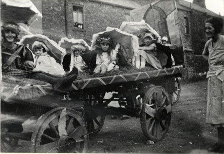 Photograph showing a cart decorated with ribbons and a tailboard; six girls aged between ten and three years are sitting on the cart wearing light dresses and headdresses and sitting under parasols; a woman is watching at the right of the picture and people can be seen indistinctly looking out of a window in the house behind the cart; the picture has been identified as depicting a carnival in Thornley