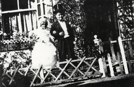 Photograph of a girl and boy, aged approximately six years dressed as a bride and groom in white frock and veil and top hat and morning suit; they are standing in front of a house with a small fence in front of them; they have been described as taking part in a carnival at Thornley