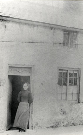 Photograph of a woman wearing a dark high-necked blouse and long dark skirt, standing in the doorway of a house with a window on the right and a window on the first floor; she has been identified as Mrs. Blunt in Quarry Street, Thornley