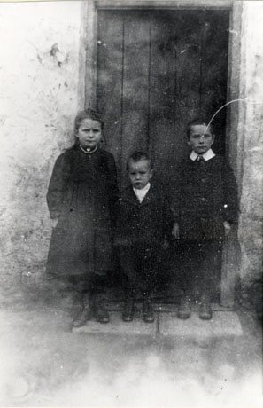 Photograph showing a girl aged approximately eight years standing with a boy aged approximately five years and a boy aged approximately seven years; the girl is wearing a dark overcoat and boots and the two boys dark suits and boots; they are standing in row in the doorway of a house; they have been identified as being in Thornley