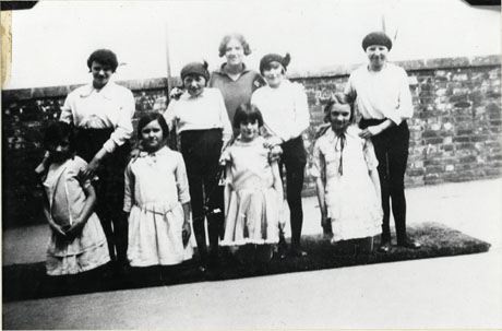 Photograph showing four girls aged approximately ten years kneeling on a mat in a yard with wall behind; the girls are wearing light dresses and four girls aged approximately fifteen, wearing blouses and dark tights are standing behind them on the mat; a woman is standing behind the group; the photograph has been described as Thornley Council School, Physical Training Display, June 1930