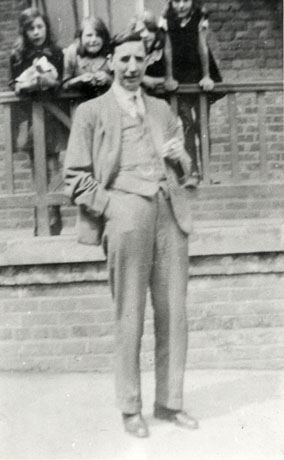 Photograph of a man standing in front of a walkway on which four girls aged approximately ten years are standing watching; he is dressed in a suit and tie and has a cigarette in his left hand; he has been identified as the Head Master of Thornley Council School at the physical training display