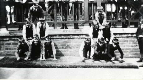 Photograph of twelve boys, aged approximately eleven years, in two pyramids of six each on a mat against a raised walkway; the legs of spectators on the walkway and part of a boy at the right of the picture can be seen; the photograph has been identified as depicting the physical training display at Thornley Council School