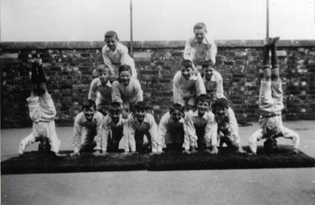 Photograph of twelve boys, aged approximately eleven years, in two pyramids of six each on a mat in a yard in front of a brick wall; a boy at either end of the mat is standing on his head; they have been identified as taking part in the physical training display at Thornton Council School