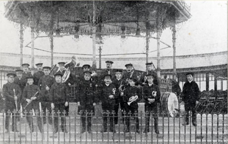 Photograph of nineteen men in uniform, carrying musical instruments, standing on the steps of a bandstand; the elaborate roof of the bandstand and a small fence surrounding it can be seen; they have been identified as members of the band of the Salvation Army in Thornley, 1916
