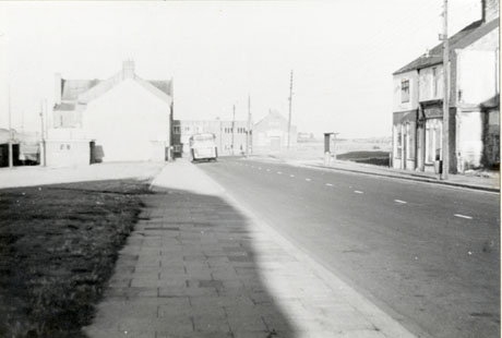 Photograph showing a road running away from the camera with an open space on the left of the road; the end of a building can be seen on the left where a bus is parked, which has been identified as the workmen's club; a building can be seen at the end of the road which has been identified as the community centre; two shops can be seen on the right; the rest of the buildings have possibly been demolished; the road has been identified as Hartlepool Street, Thornley