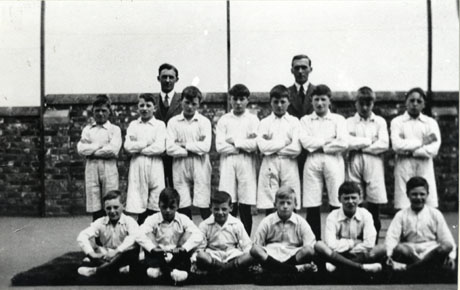 Photograph showing eight boys aged approximately twelve years dressed in white shirts and shorts standing in a row behind six boys similarly dressed sitting on a mat; behind the boys are two men and a wall; they have been identified as being part of the physical training display at Thornley Council School