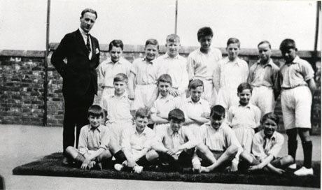 Photograph of seven boys, dressed in white shirts and shorts, standing behind nine boys, similarly dressed, sitting on a mat; the boys are aged approximately ten years and are accompanied by a man; a wall is behind the group; they have been identified as being part of the Physical Training Display at Thornley Council School