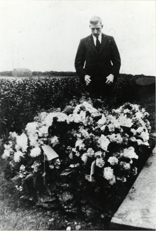Photograph of a man wearing a dark suit and tie and holding his hat in his hands standing in front of a low hedge and looking at a pile of funeral flowers; the photograph has been identified as Mrs. Gair's Funeral, Thornley New Cemetery