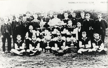 Postcard photograph, entitled Thornley Football Club, showing twelve men in football strip, accompanied by nineteen other men; in front of the group are two large trophy cups; the group is photographed against trees