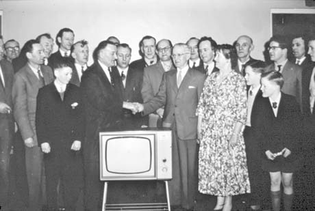 Photograph of a group of eighteen men standing against a wall, watching a man shaking hands with a man who is standing next to a woman in a flowered dress; in front of the men shaking hands is a television set; three boys, aged approximately between ten and twelve years, are standing at the front of the group watching; the photograph has been identified as Golden Wedding Presentation, Thornley