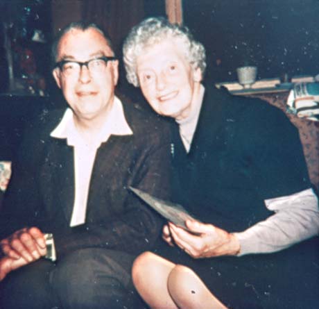 Photograph of a middle-aged man wearing a jacket, open-necked shirt and trousers, sitting next to a woman wering a pinafore dress with a jumper underneath it; the woman is holding what may be a card; they are sitting on a sofa and an uncurtained window with darkness outside can be seen behind them; they have been identified as Vicar and Mrs. Mould
