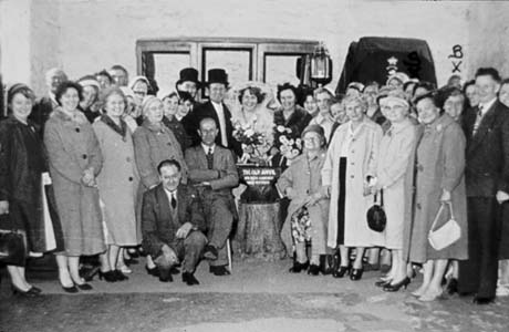 Photograph of approximately thirty middle-aged people standing either side of a couple dressed as bride and groom and an anvil on a stand with the words The Old Anvil on it; the people have been identified as members of the Over Twenty Club of Thornley on a trip to Gretna Green