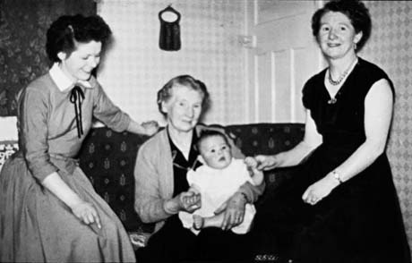 Photograph of an elderly woman sitting on a sofa holding a baby; on her right is a young woman and on her left a middle-aged woman; behind her the walls and door of living room can be seen; the elderly woman has been identified as Mrs. Jonas Gott with three other generations of her family