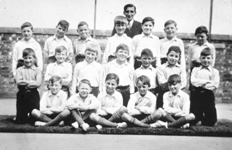 Photograph of eighteen boys, aged approximately ten years and wearing shirts, dark trousers, socks and plimsolls, posed in three rows on a mat in front of a wall with a man behind them; they have been identified as taking part in the Physical Training Display at Thornley Council School