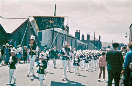 Photograph showing two girls aged approximately sixteen years holding a banner reading Peterlee Gold Star 1964 Jazz Band walking along a street followed by a procession of children aged approximately seven years in uniform; groups of people can be seen on either side of the street watching the parade; the photograph has been identified as Carnival, Thornley