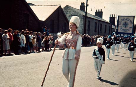 Photograph of a girl aged approximately sixteen years dressed in the uniform of a jazz band and carrying a ceremonial stick; she is followed by a procession of children, aged approximately ten years, in uniform, carrying a banner reading: Blue Stars Jazz Band Horden; the procession is walking along a street in Thornley with crowds watching during a Carnival