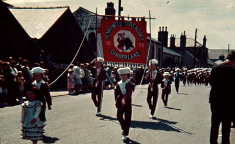 Photograph of children aged approximately between six and twelve years in the uniform of a jazz band walking along a street in Thornley as part of a Carnival; they are carrying a banner reading: Pennywell Toreadors Sunderland 1963; groups of people watching the procession can be seen