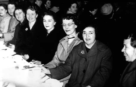 Photograph of eight young women wearing overcoats sitting at a table laid with a cloth and cutlery; they have been identified as Mothers' Club Party in Thornley