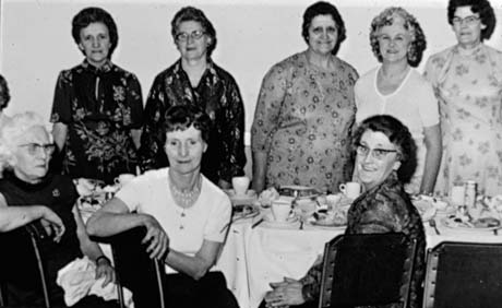 Photograph showing six women wearing dresses standing behind a table on which plates of food and cups and saucers can be seen; three women are sitting in front of the table; the photograph has been identified as Mothers' Club 25th Anniversary, Thornley