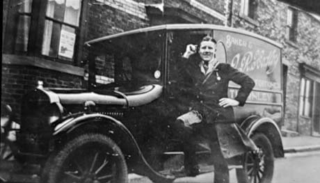 Photograph showing a motor van standing in a road in front of terraced houses; on the side of the van are the following words: I. R. Hedley, Baker and Confectioner, Thornley; a young man wearing a dark jacket over a dark apron is standing with one foot on the running board of the van; the photograph ahs been described as Hedley The Baker
