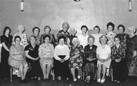 Photograph of eleven women in formal dresses, a number of which are full-length, standing against a wall; in front of them are eight other women, similarly dressed, sitting on chairs; the group has been described as Mothers' Club Committee, Thornley