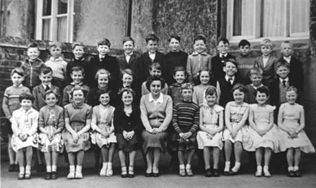 Photograph of thirty three children, aged approximately seven years, grouped against the wall of a building; a woman is sitting with the children on the front row; the group has been identified as pupils and their teacher at the Junior School at Thornley