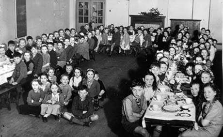 Photograph showing approximately seventy eight children sitting round three long tables arranged as three sides of an oblong in a hall; tea cups, cakes and plates can be seen on the tables; the photograph has been identified as recording a party at the Junior School at Thornley