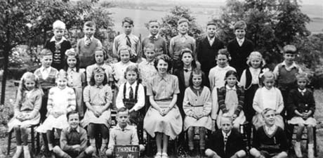 Photograph showing twenty eight children, aged approximately seven years, grouped in front of bushes with fields in the distance; a woman, presumably their teacher, is sitting on the front row; a child on the front row is holding a notice, of which only the word, Thornley, can be read; the children have ben identified as pupils at Thornley Junior School