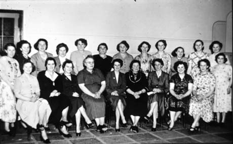 Photograph showing fourteen women wearing dresses standing against a wall with ten women wearing dresses sitting in front of them; the group has been identified as Mothers' Club in Workingmens' Club, Thornley