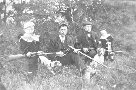 Photograph showing, on the left of the picture, a boy aged approximately seven years, wearing a suit and cap, holding an air rifle; next to him is a dog and, next to the dog, a man wearing a suit, tie and cap, sitting on the ground and holding an air rifle, with the body of a rabbit lying on his foot; this man and boy have been identified as Tom Armstrong and son; next to Tom Armstrong is another man wearing a suit, tie and cap, carrying an air rifle and with a dog sitting in front of him; next to him at the right of the picture is a small boy, aged approximately five years, holding a dead rabbit; this man and boy have ben identified as Chas Bullock and son; they are described as being in Thornley