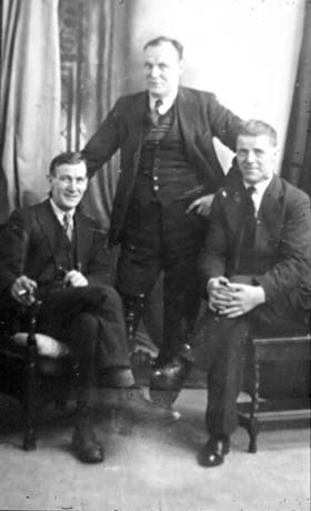 Photograph showing a man wearing a suit and tie and with a cigarette in his hand, sitting in a chair at the left of the photograph; next to him is a man standing, similarly dressed; no the right of the picture is a third man also dressed in suit and tie, sitting on a chair; they have been identified as Matt, Phil, and Jack Rutherford