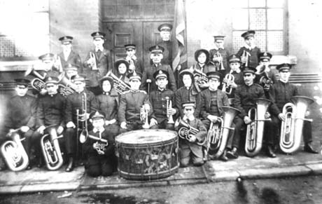 Photograph showing twenty five people dressed in uniform holding musical instruments grouped outside a building with a large door and two windows; they have been identified as members of the Salvation Army Band in Thornley