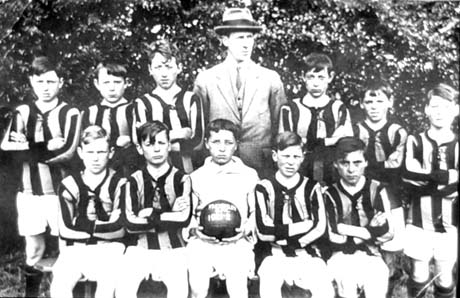 Photograph showing eleven boys in football strip posed in front of bushes, with a man wearing a suit, tie and Trilby hat; a boy on the front row is holding a football, on which illegible words are written; the boys have been identified as members of a school football team in Thornley; the man has been identified as Mr. Wilkinson, a school teacher; the boy holding the ball, the goal keeper, has been identified as Bob Bullock