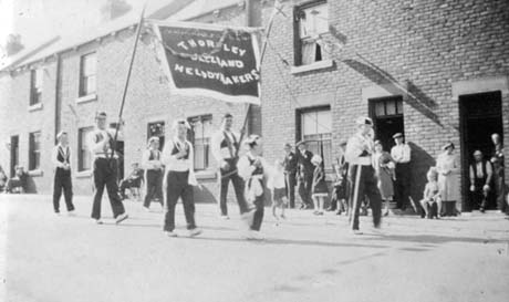 Photograph showing five boys and two men carrying a banner reading Thornley Jazz Band Melody Makers walking along a street with terraced houses behind them; approximately fifteen people are watching the banner carriers; the street has been identified as High Street, Thornley