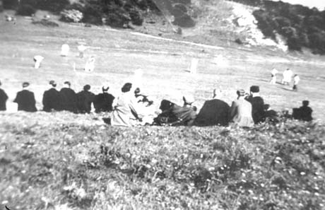 Photograph showing the backs of approximately fifteen people watching a cricket match being played on an open space with a hillside beyond it; the figures of the cricketers are difficult to discern; the photograph has been identified as Cricket Match in The Hilly Before It Was Filled In in Thornley