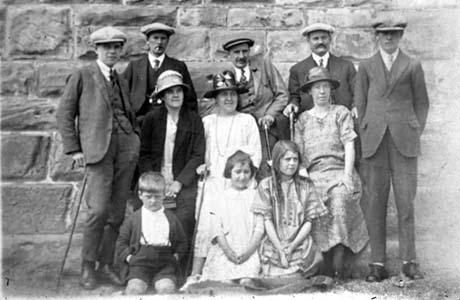 Photograph showing five men dressed in suits, ties and caps; three women, dressed in dresses and hats; two girls, aged approximately between eight and ten years, dressed in frocks; and a boy, aged approximately seven years, dressed in a jacket and short trousers; all are posed against a stone wall; they have been described as Willy and Bosomworth Families of Thornley