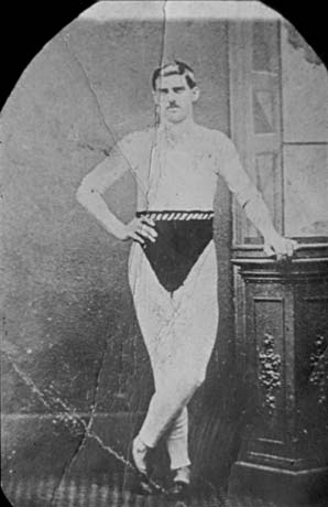Photograph of a man wearing long johns with a dark pair of trunks over them; he is standing leaning with his left hand on a piece of furniture, possibly in a photographer's studio; he has been identified as Mr. Wallis, of Thornley, a runner