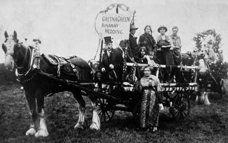 Photograph showing a horse harnessed to cart, on which four people can be seen standing and three people sitting; on the front of the cart is a sign reading Gretna Green Runaway Wedding; a man in a top hat is sitting on the cart holding the reins and a blurred figure is standing on the ground near the front of the cart; the cart has been identified as taking part in the First Carnival at Thornley