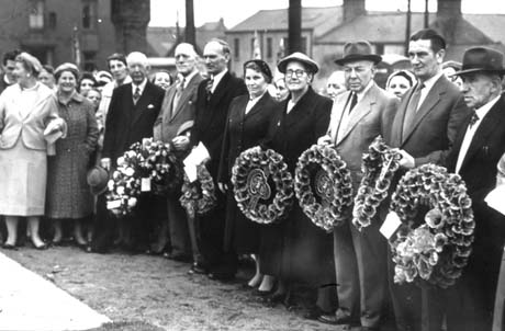 Photograph showing a crowd of approximately thirty people with the tops of houses behind them; on the front row of the crowd are five middle-aged women and six middle-aged men, six of whom are holding wreaths of poppies; the crowd has been identified as being in Thornley