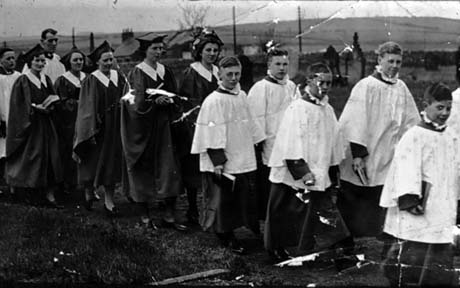 Photograph showing seven boys wearing cassocks and surplices and five young women wearing academic gowns and academic hats, walking in a procession through a graveyard with fields in the background; they have been described as the choir in procession in Thornley