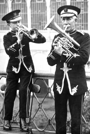 Photograph showing a boy, aged approximately eleven years, standing on a chair wearing a uniform and blowing a trumpet; on the right of the picture is an elderly man, also in uniform, also blowing a trumpet; behind them is an indistinct building; the boy has been identified as Edward Kitto and the man as Mr. Johnson