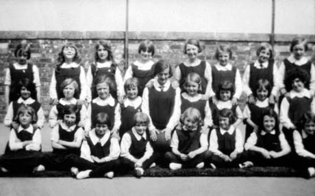 Photograph showing twenty four girls, aged approximately eleven years, all wearing gym slips, posed against a brick wall; an older girl is posed in the middle of the group; they have been described as taking part in a physical training display at Thornley