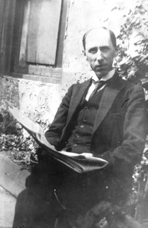 Photograph showing a man wearing a suit, waistcoat and wing collar, sitting with his back to the wall of a building and holding a newspaper in his hand; he has been identified as Albert H. Oswald, composer and organist, in Thornley