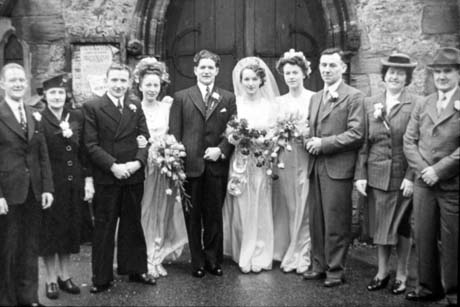 Photograph showing a newly-married bride and groom with a bridesmaid on either side of the couple; on either side of the bride, groom and bridesmaids are two men and a woman with button holes in their dress; the photograph has been identified as Kitto's Wedding Group, Thornley; the group is standing in front of the west door of a church